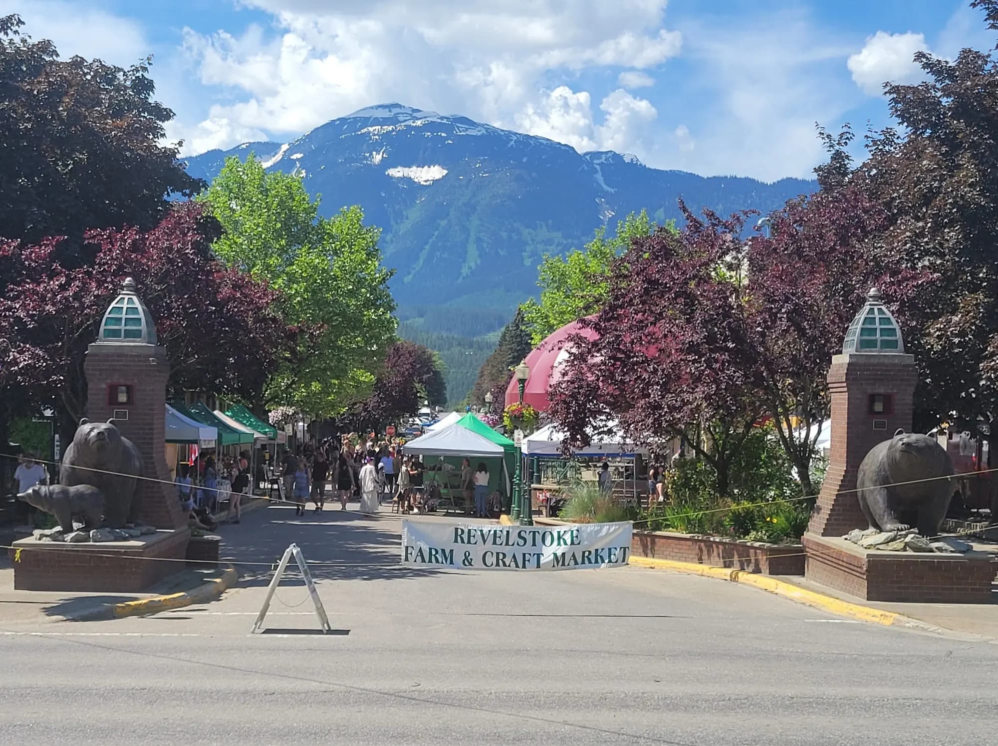 Grizzly Plaza on a Saturday — market tents lining the street under Mount Mackenzie, flanked by the plaza's stone bear pillars